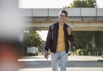 Lächelnder junger Mann, der beim Gehen auf der Straße ein Smartphone benutzt - UUF20918