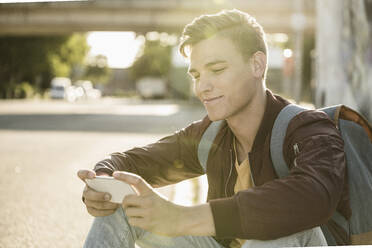 Lächelnder gut aussehender Mann, der ein Smartphone benutzt, während er an einem sonnigen Tag auf der Straße sitzt - UUF20926