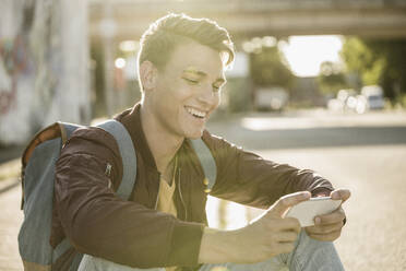 Glücklicher junger Mann, der sein Smartphone benutzt, während er an einem sonnigen Tag auf der Straße sitzt - UUF20927