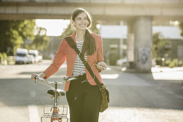Lächelnde Frau mit Fahrrad hört Musik über ihr Smartphone, während sie auf der Straße geht - UUF20929