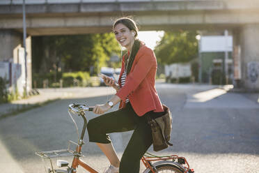 Happy young woman using smart phone while riding bicycle on street in city - UUF20937
