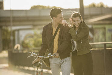 Happy couple with bicycle walking on street during sunny day - UUF20944