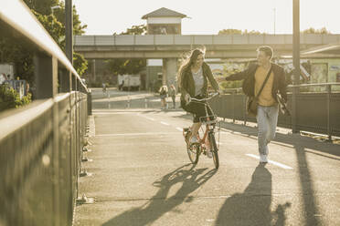 Glücklicher junger Mann, der an einer Freundin vorbeiläuft, die mit dem Fahrrad in der Stadt unterwegs ist, an einem sonnigen Tag - UUF20945