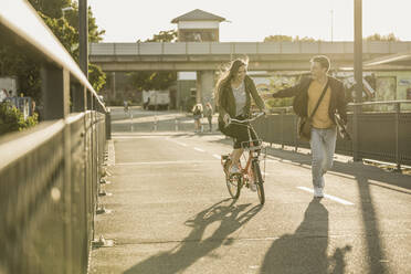 Happy young man running by girlfriend riding bicycle in city during sunny day - UUF20945