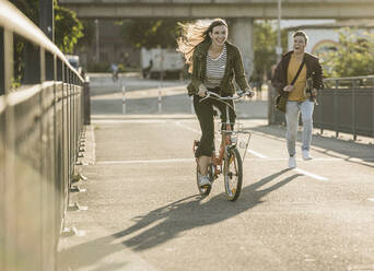 Freund läuft hinter einer glücklichen Freundin, die in der Stadt an einem sonnigen Tag Fahrrad fährt - UUF20946