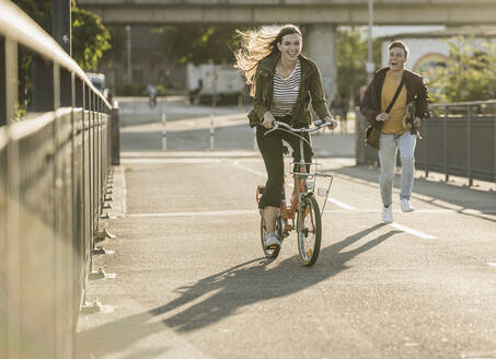 Freund läuft hinter einer glücklichen Freundin, die in der Stadt an einem sonnigen Tag Fahrrad fährt - UUF20946