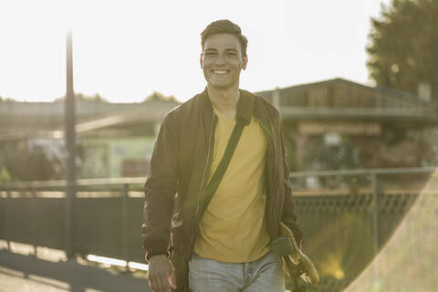 Smiling young man carrying skateboard while standing in city during sunny day stock photo
