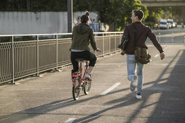 Junger Mann, der ein Skateboard trägt, während er an seiner Freundin vorbeirennt, die in der Stadt Fahrrad fährt - UUF20950