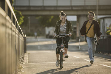 Fröhlicher Freund läuft hinter seiner Freundin auf dem Fahrrad in der Stadt an einem sonnigen Tag - UUF20951
