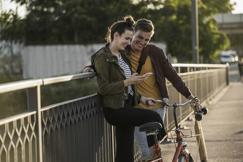 Lächelndes junges Paar schaut auf sein Smartphone, während es mit dem Fahrrad in der Stadt steht - UUF20952