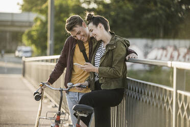 Smiling boyfriend and girlfriend looking at smart phone while standing with bicycle in city - UUF20953