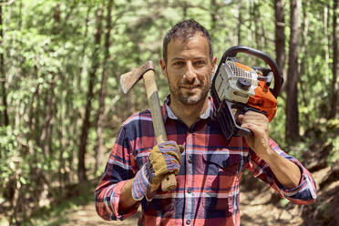 Lächelnder Holzfäller mit Axt und Kettensäge im Wald stehend - VEGF02780