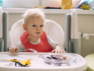 Baby boy sitting on high chair while eating breakfast in kitchen - KNTF05188
