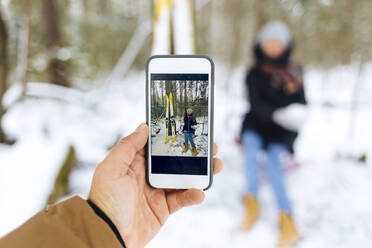 Man photographing girlfriend by skis through smart phone in forest during winter - KNTF05198