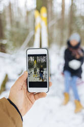 Boyfriend photographing girlfriend by skis through mobile phone in forest during winter - KNTF05199
