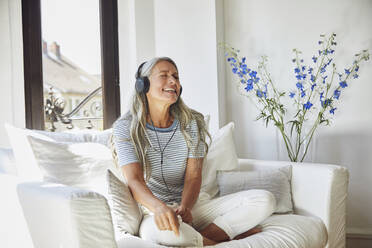 Happy woman listening to music through headphones on sofa at home - MCF01134