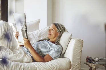 Woman reading magazine while lying on sofa in living room - MCF01149