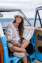 Stylish smiling lady in hat with bottle looking at camera with smile while opening door of car on beach - ADSF11214