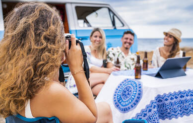 Fröhliche Freundin, die lächelt und eine Getränkeflasche in der Hand hält, sitzt am Tisch, während eine langhaarige Frau in der Nähe eines blauen Autos ein Foto macht - ADSF11216