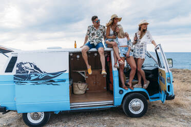 Fröhliche Gruppe junger Leute mit Flaschenbindung auf dem Dach eines hellen Minivans am Strand in einem sonnigen Tag - ADSF11224