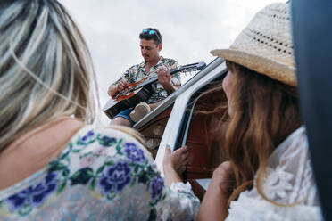 Enthusiastischer Mann spielt Gitarre auf dem Autodach sitzend, während reizende Damen zuhören und Lieder im Auto am Strand genießen - ADSF11227