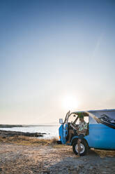 Side view of attentive adult male in tropical shirt and denim shorts focusing on map while sitting in parked blue van with opened door and creating route on beach - ADSF11236