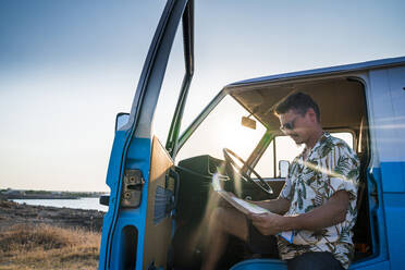 Seitenansicht eines aufmerksamen erwachsenen Mannes in Tropenhemd und Jeansshorts, der sich auf eine Landkarte konzentriert, während er in einem geparkten blauen Van mit geöffneter Tür sitzt und eine Route am Strand erstellt - ADSF11237
