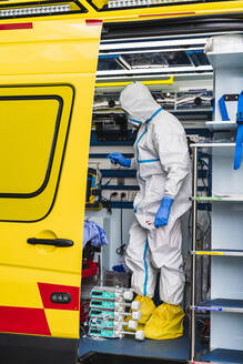 Side view of anonymous doctor in protective suit standing in ambulance car near opened door with equipment and examining patient with virus infection - ADSF11239