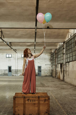 Frau steht auf Holzkiste und hält bunte Heliumballons, lizenzfreies Stockfoto