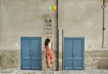 Woman climbing ladder leaning on wall while reaching for colorful helium balloons - VEGF02792