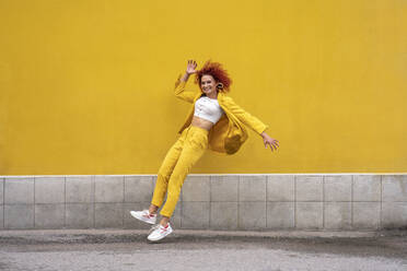 Energetic young woman in yellow suit running and jumping in front of yellow wall - VPIF02783