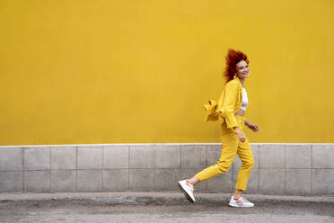 Energetic young woman in yellow suit running and jumping in front of yellow wall - VPIF02785