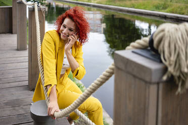 Young woman with curly hair and yellow suit sitting by the riverside, talking on ghe phone - VPIF02816
