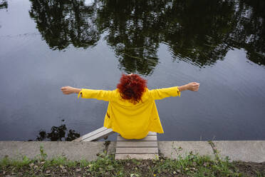 Junge Frau mit lockigem Haar und gelbem Anzug sitzt mit ausgestreckten Armen am Flussufer - VPIF02818