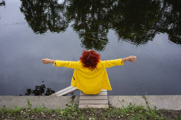 Young woman with curly hair and yellow suit sitting by the riverside with arms outstretched - VPIF02818