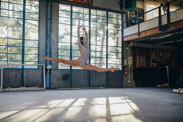 Seitenansicht einer jungen schlanken Ballerina im Gymnastikanzug, die beim Üben von Tanzbewegungen in einem hellen geräumigen Studio mit großem Fenster springt - ADSF11325