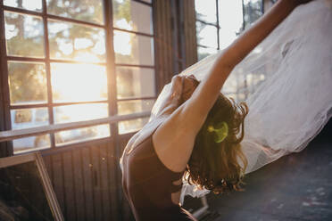 Side view of unrecognizable young dramatic female dancer in black bodysuit performing sensual dance with tulle while training alone in light spacious studio - ADSF11329