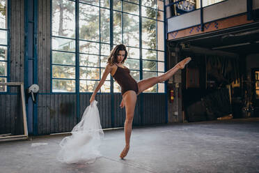 Full body of slim graceful female dancer looking at camera in black bodysuit and pointe shoes performing dance with transparent light tulle during rehearsal in studio - ADSF11330