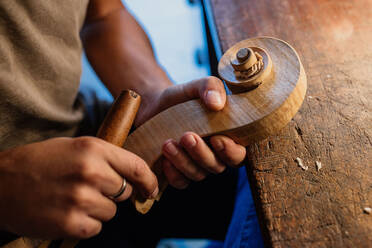 Cropped faceless craftsman carving swirl of violin head while working with wooden detail using bit at table in workshop - ADSF11332