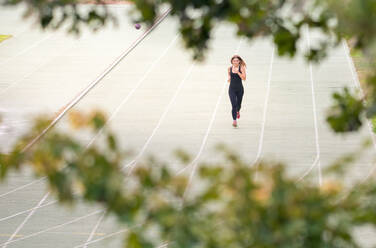 View between trees of a young fit woman running in a track outdoors in a sunny day - ADSF11337