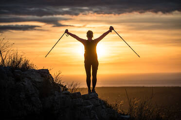 Silhouette of unrecognizable woman standing with walking sticks on stony hill spreading arms wide in sunset - ADSF11339