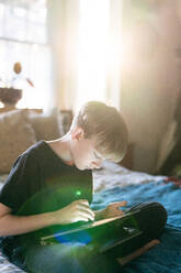 Jugendlicher auf dem Bett sitzend mit Stift und 2in1-Laptop in der Abenddämmerung - CAVF88427