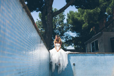 Blick von unten auf eine junge verträumte Frau in einem eleganten weißen Hochzeitskleid, die auf dem Rand eines leeren Swimmingpools in der Nähe eines grünen Baumes ruht und verträumt in die Ferne schaut - ADSF11383