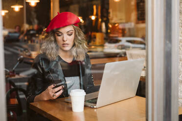 Trendige junge Frau mit roter Baskenmütze telefoniert mit ihrem Handy, während sie mit ihrem Laptop in einem Restaurant am Tisch sitzt - ADSF11398