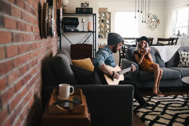 Männlicher Gitarrist in Freizeitkleidung spielt Akustikgitarre, während er auf dem Sofa neben seiner Partnerin mit einer Tasse Heißgetränk in einer Wohnung sitzt - ADSF11402