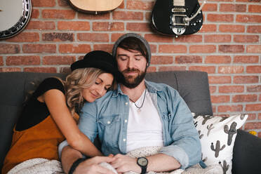 Lächelnder Freund in Jeansjacke, der seine Freundin mit Hut umarmt, während er mit geschlossenen Augen auf einer gemütlichen Couch sitzt, die mit einer Decke bedeckt ist, in der Nähe einer Backsteinmauer mit Musikinstrumenten - ADSF11403