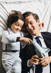 From above of cheerful boy in astronaut costume and dad in formal suit relaxing together in bedroom and playing with toy rockets - ADSF11406