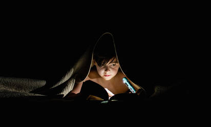 Kleiner Junge liest unter einer Decke mit einer Taschenlampe ein Buch. - CAVF88503