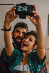 Affectionate hipster couple hugging and taking selfie with photo on camera while sitting on floor at home - ADSF11416