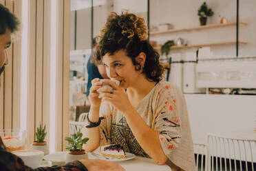Seitenansicht einer glücklichen, lächelnden Frau, die aus einem Becher nippt, während sie in einem modernen Coffeeshop sitzt - ADSF11425